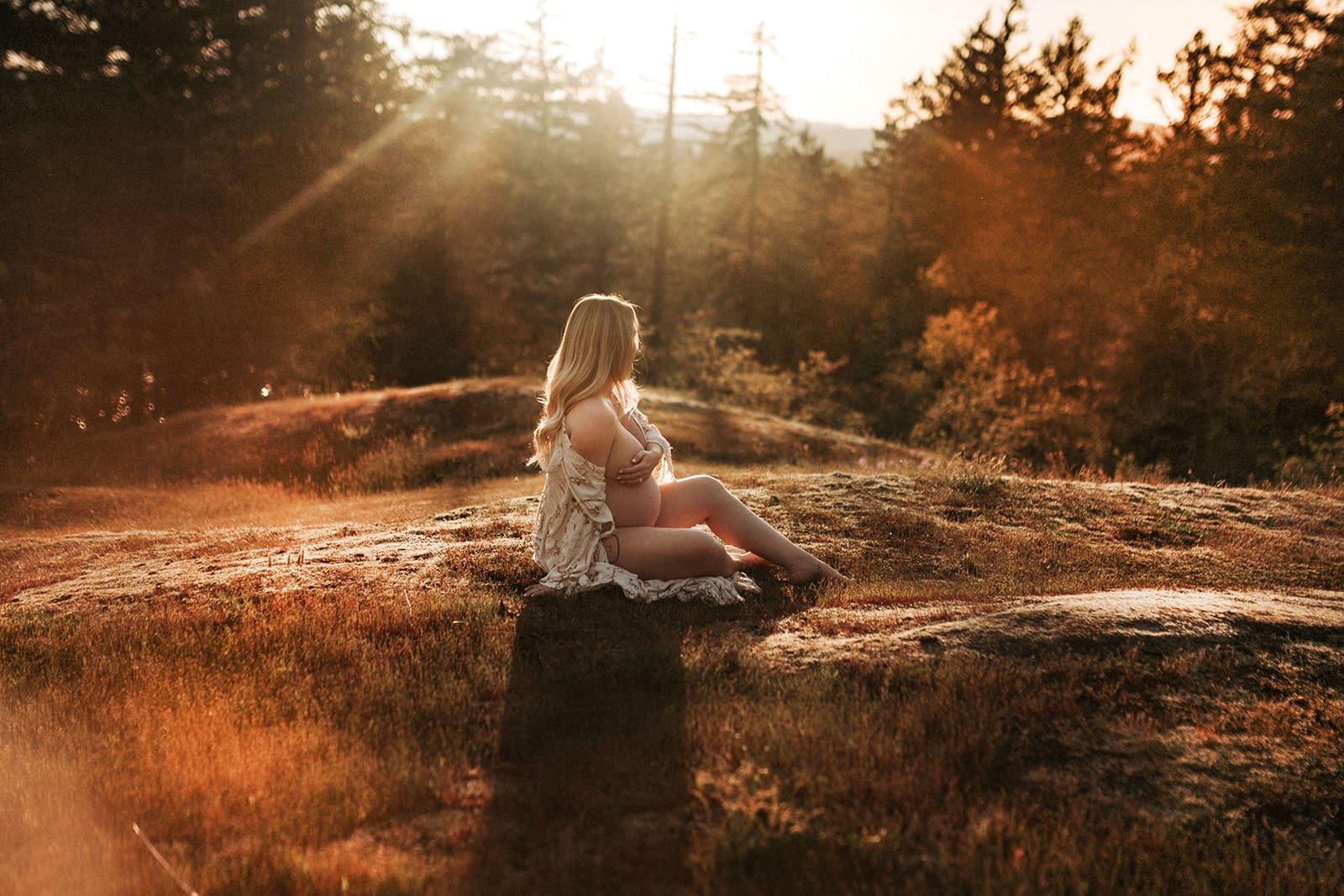 Pregnant Mom sitting on a cliff facing away from the camera, exposing her pregnant tummy at golden hour in Victoria, BC
