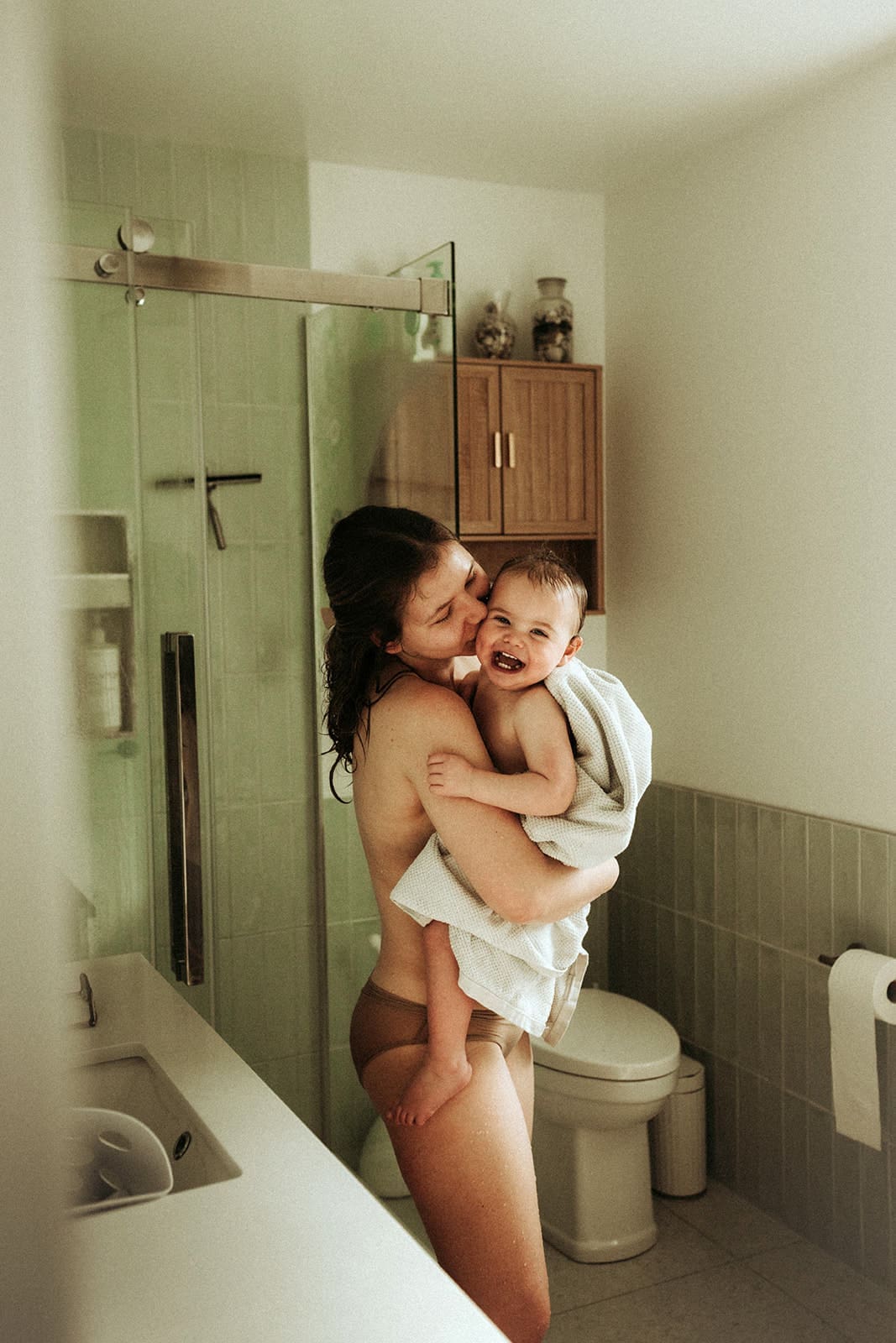 Mom and toddler having a cozy hug after bath standing in the bathroom with towel wrapped around toddler, mom kissing her cheek and her laughing