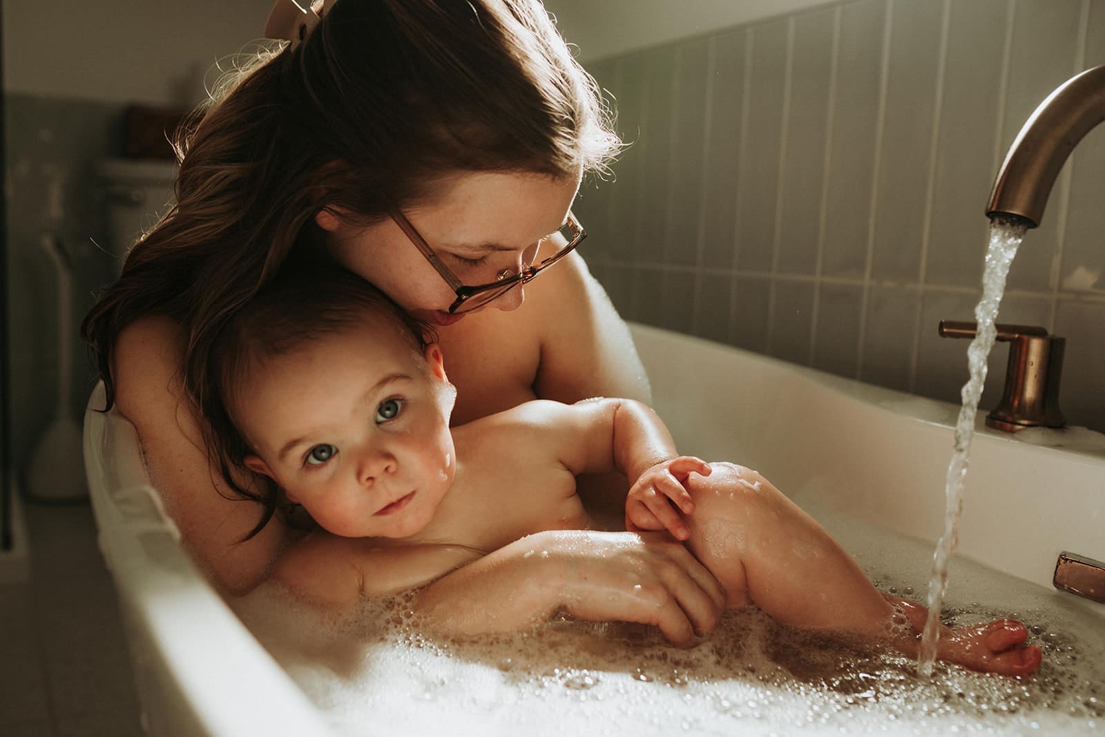 mom and daughter cuddling in tub with bubbles and the tap running