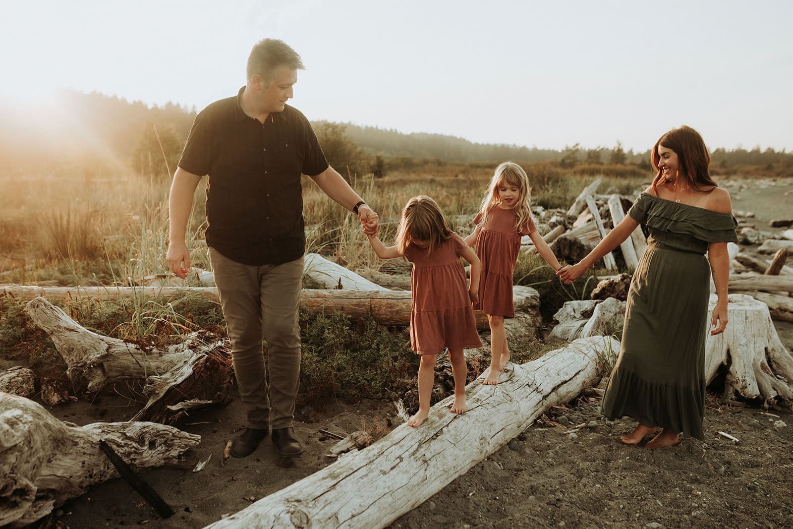 A mom and Dad holding their daughters hands as they both walk along a driftwood log trying to keep their balance